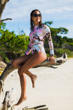 Combi surf femme rose floral UPF50+ en pied, silhouette entière portée par Moea, assise sur un tronc de bois flotté