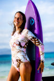 Combi surf femme manches longues rose floral UPF50+ portée par Moea avec un surf sur la plage